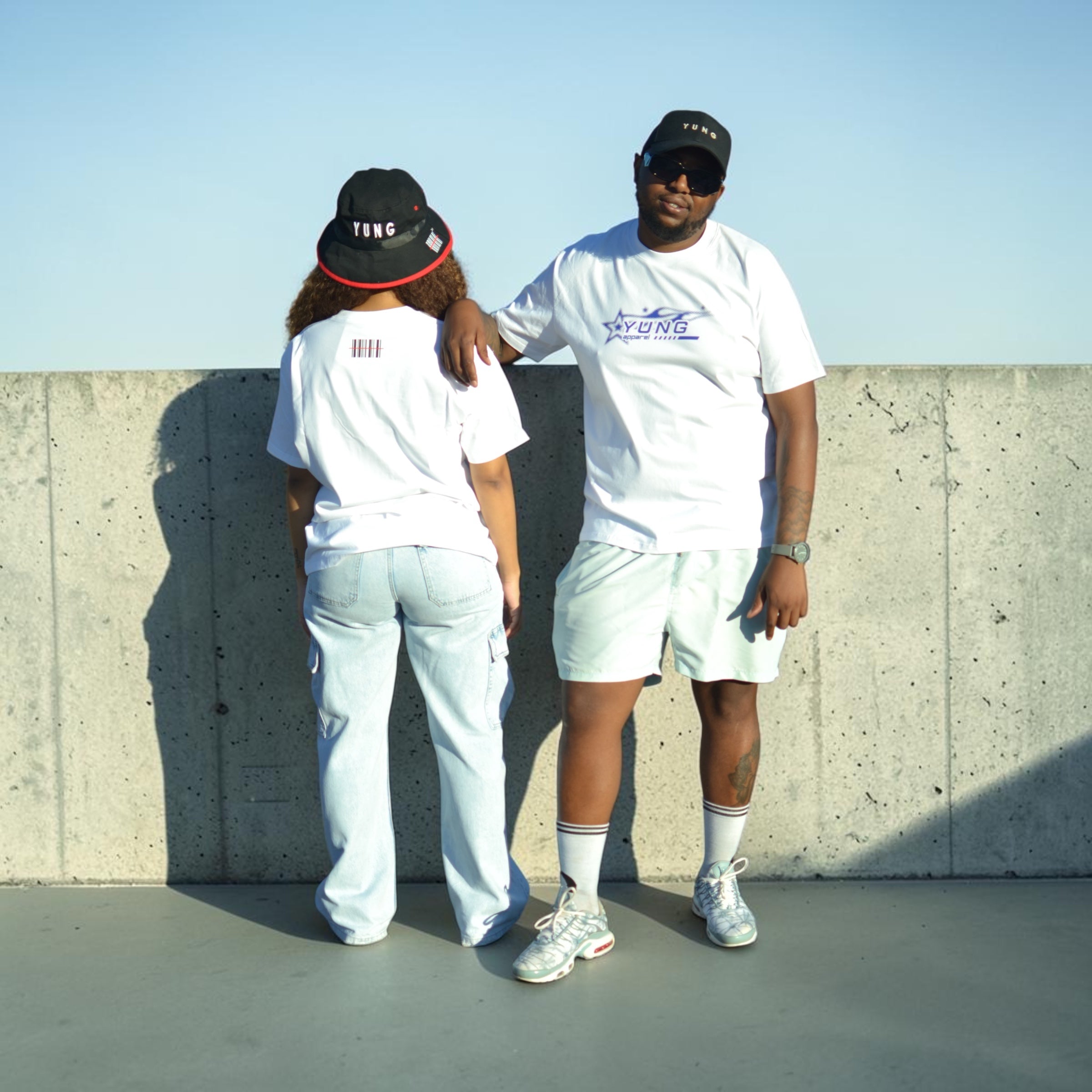Two people wearing matching white YUNG t-shirts and light-colored pants standing against a concrete wall.
South Africa