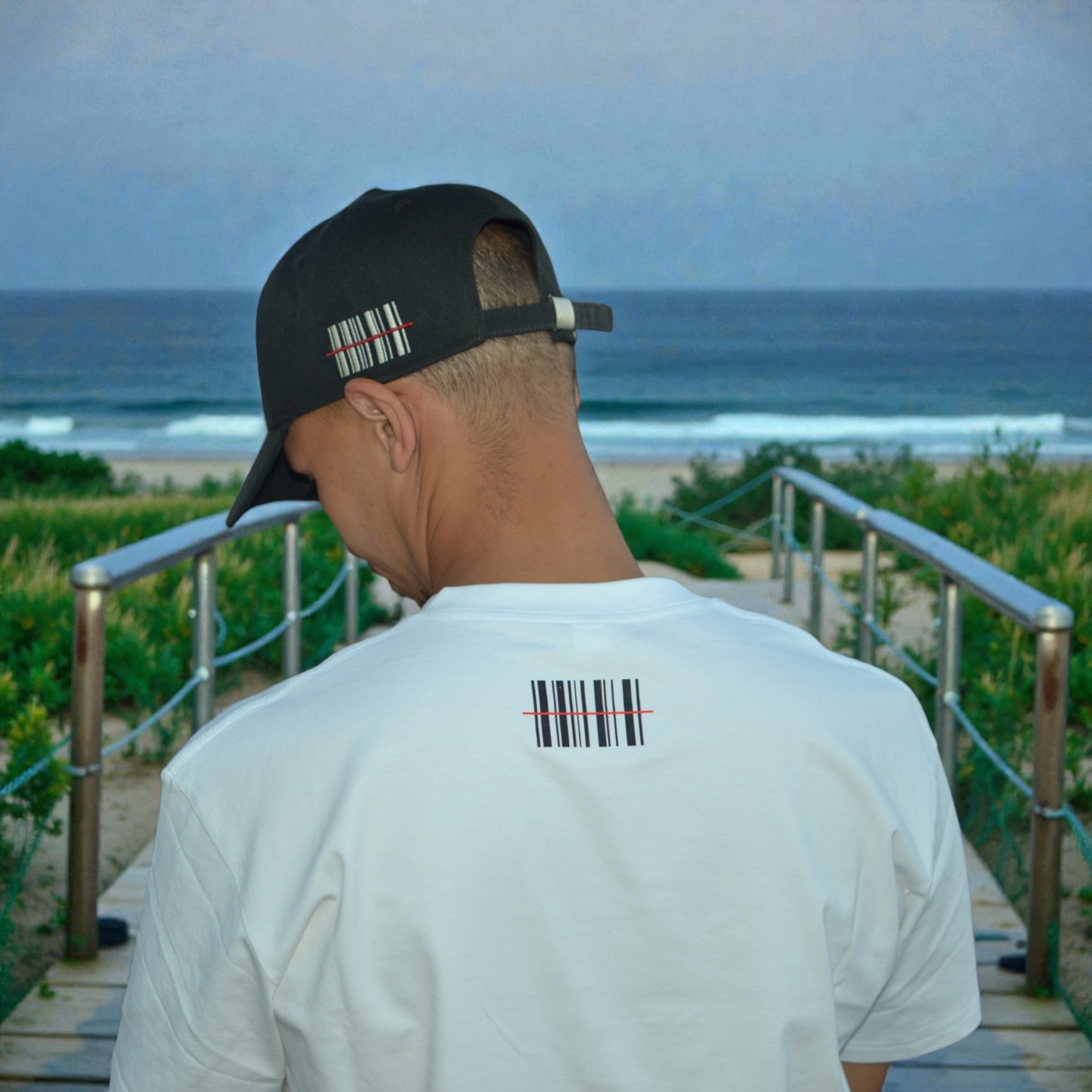 Person wearing a YUNG white t-shirt and black baseball cap with barcode designs, standing on a pathway by the beach.
South Africa