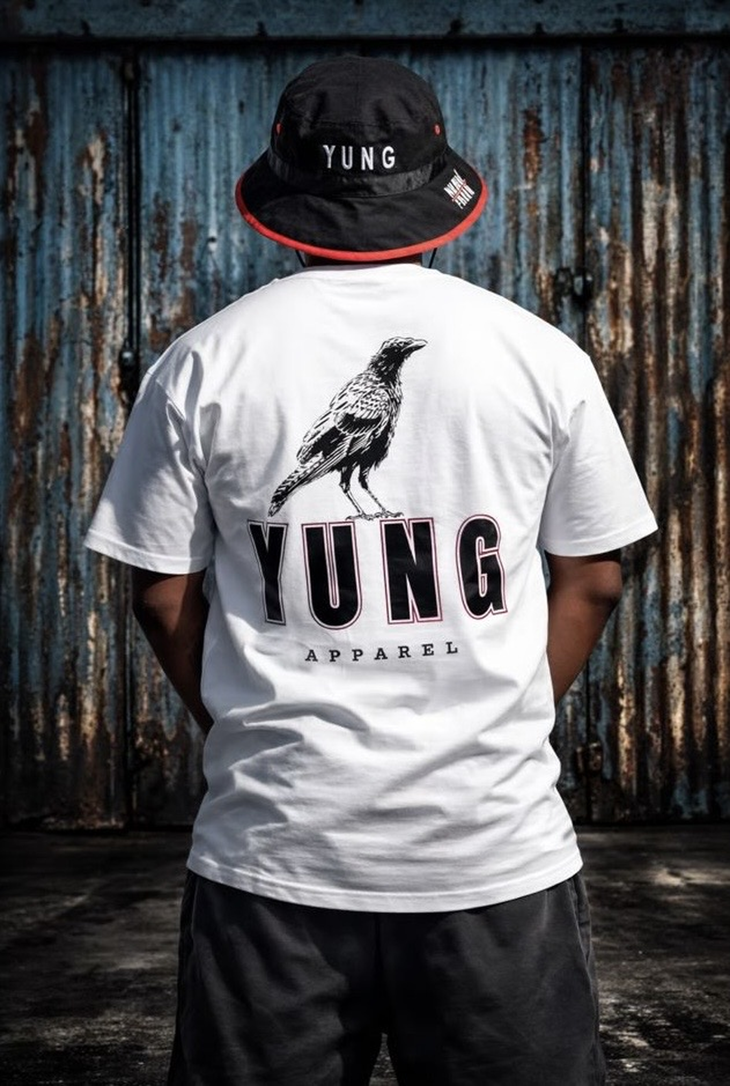 Person wearing a white 'YUNG APPAREL' t-shirt with a bird graphic and text in the back, standing against a rusty metal wall.
South Africa