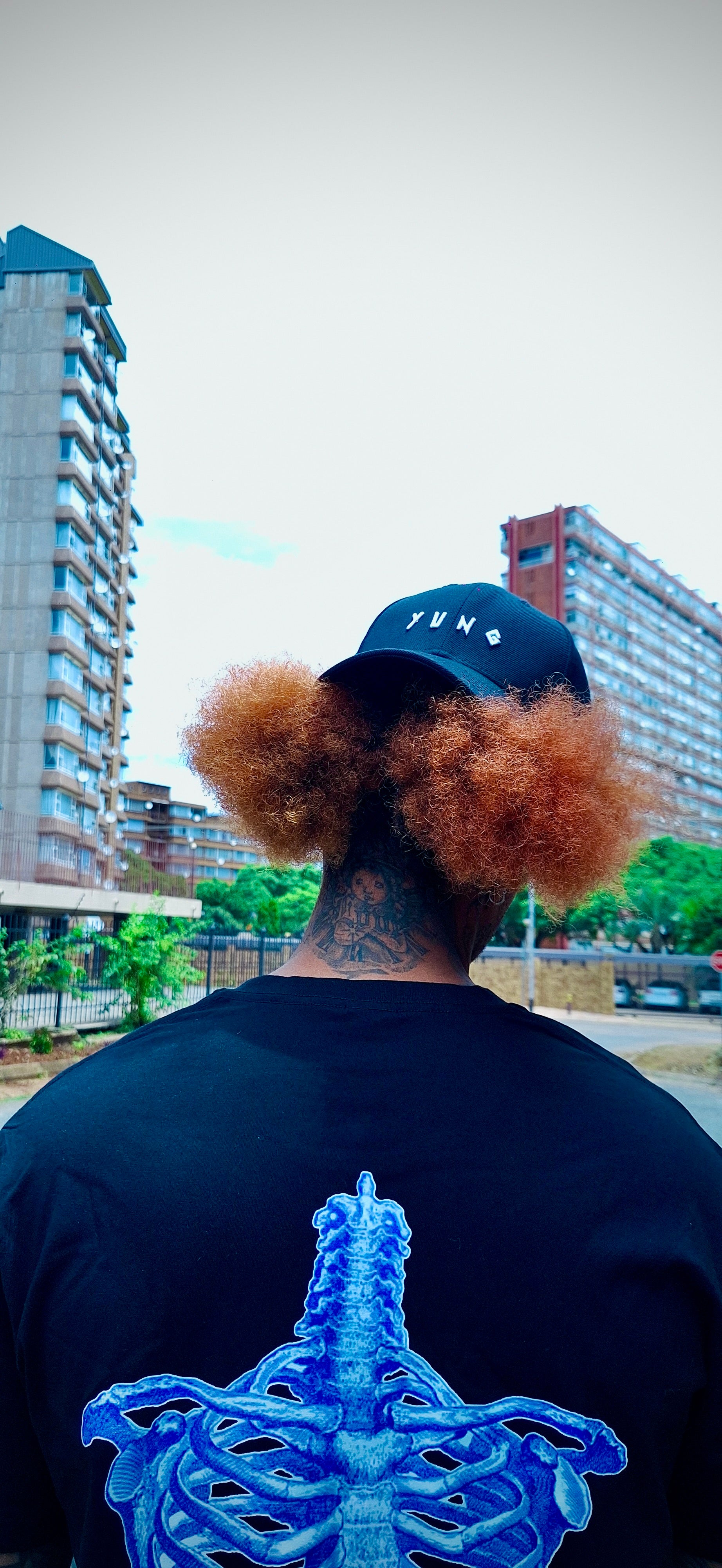 Person wearing a cap with 'YUNG' logo, back view of black shirt with YUNG blue skeleton design, urban setting
South Africa