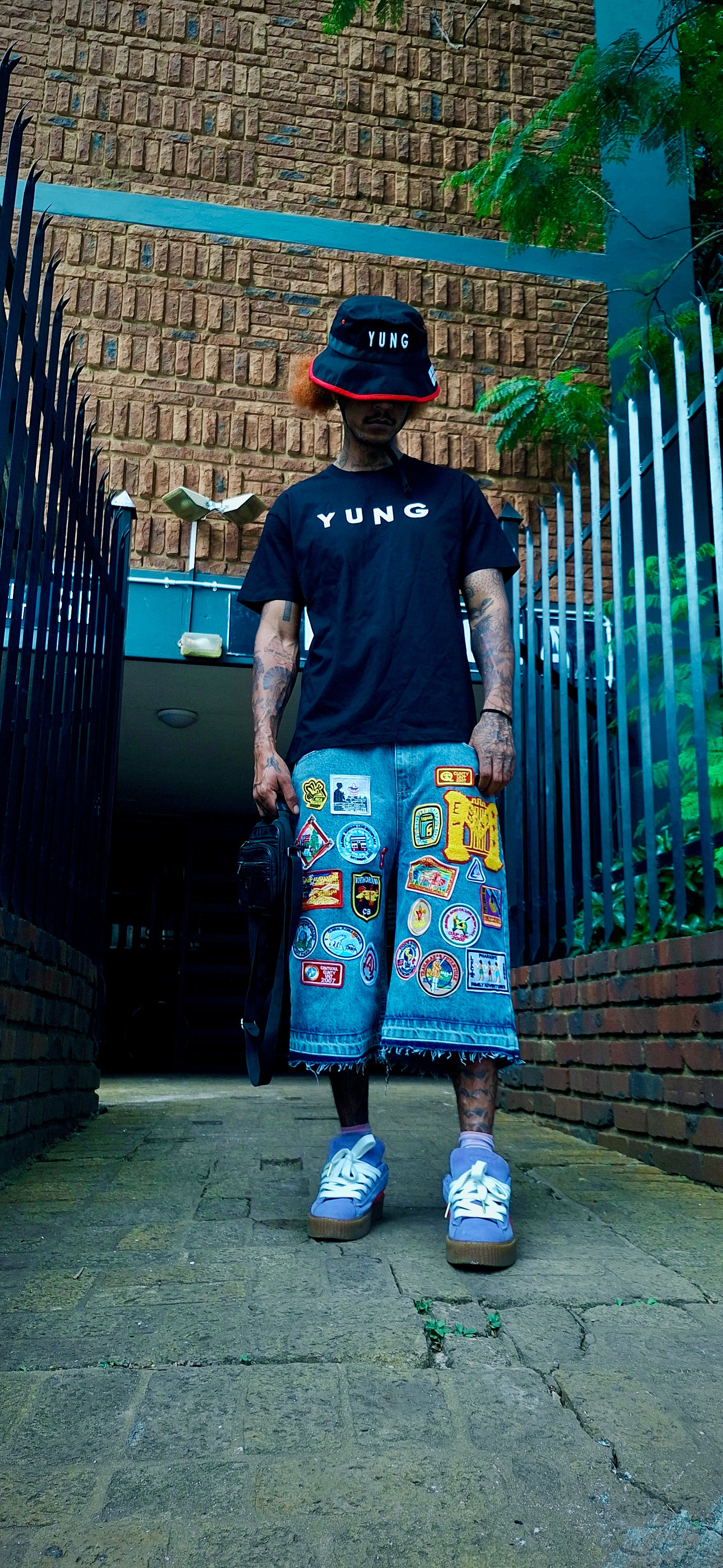 Person wearing a black 'YUNG' t-shirt, bucket hat, colorful shorts, and blue sneakers standing in front of a textured wall.
South Africa