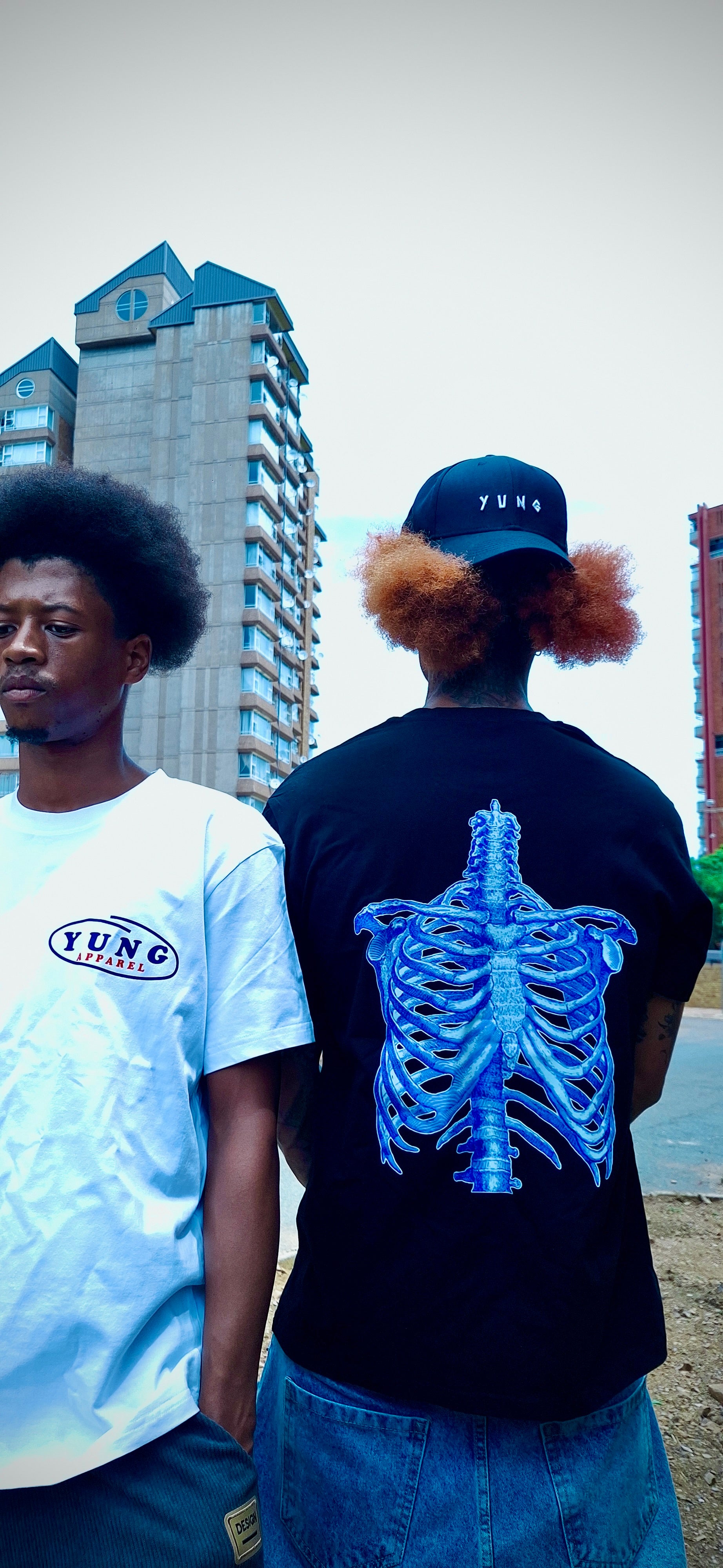 Two people standing outdoors with one wearing a YUNG black t-shirt with a blue skeleton design on the back and one person wearing a white t-shirt with 'YUNG' branding on left side of the chest in front of a burning building.
South Africa