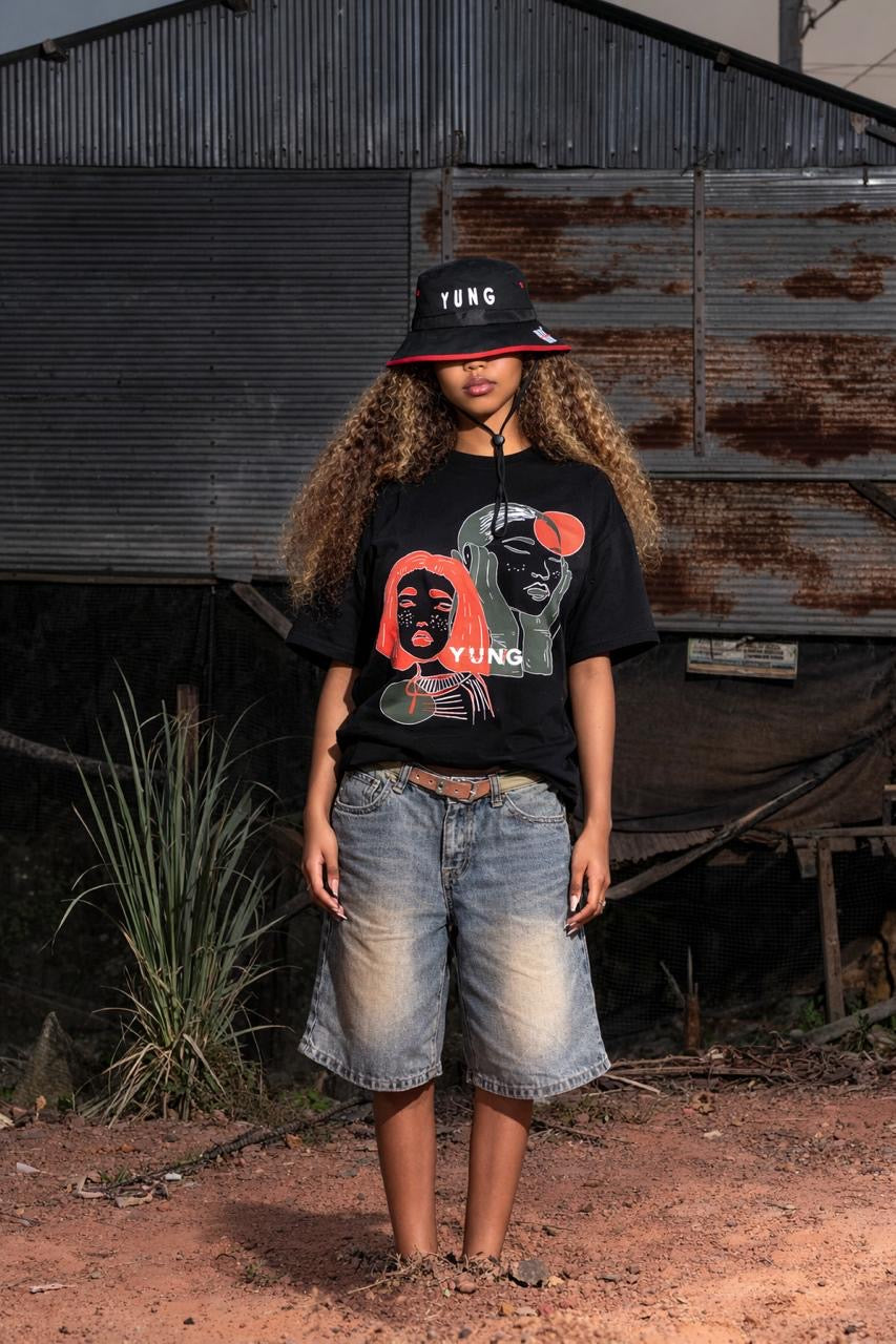 Person wearing a black Y U N G Girls Got Goals t-shirt with graphic design and denim shorts standing in front of a rustic metal wall.
South Africa