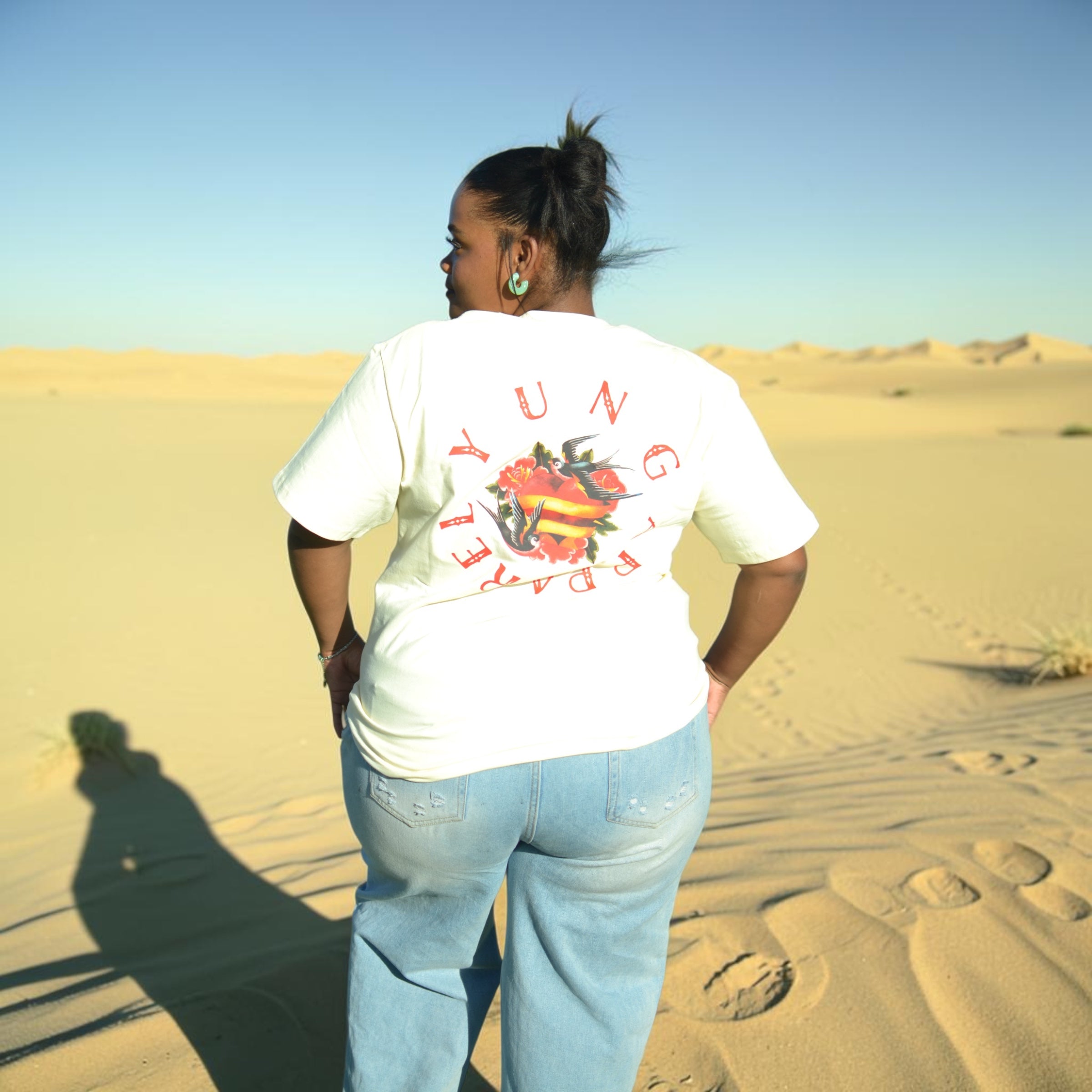 Person wearing a cream white Y U N G t-shirt with colorful graphics and text in a desert setting
South Africa