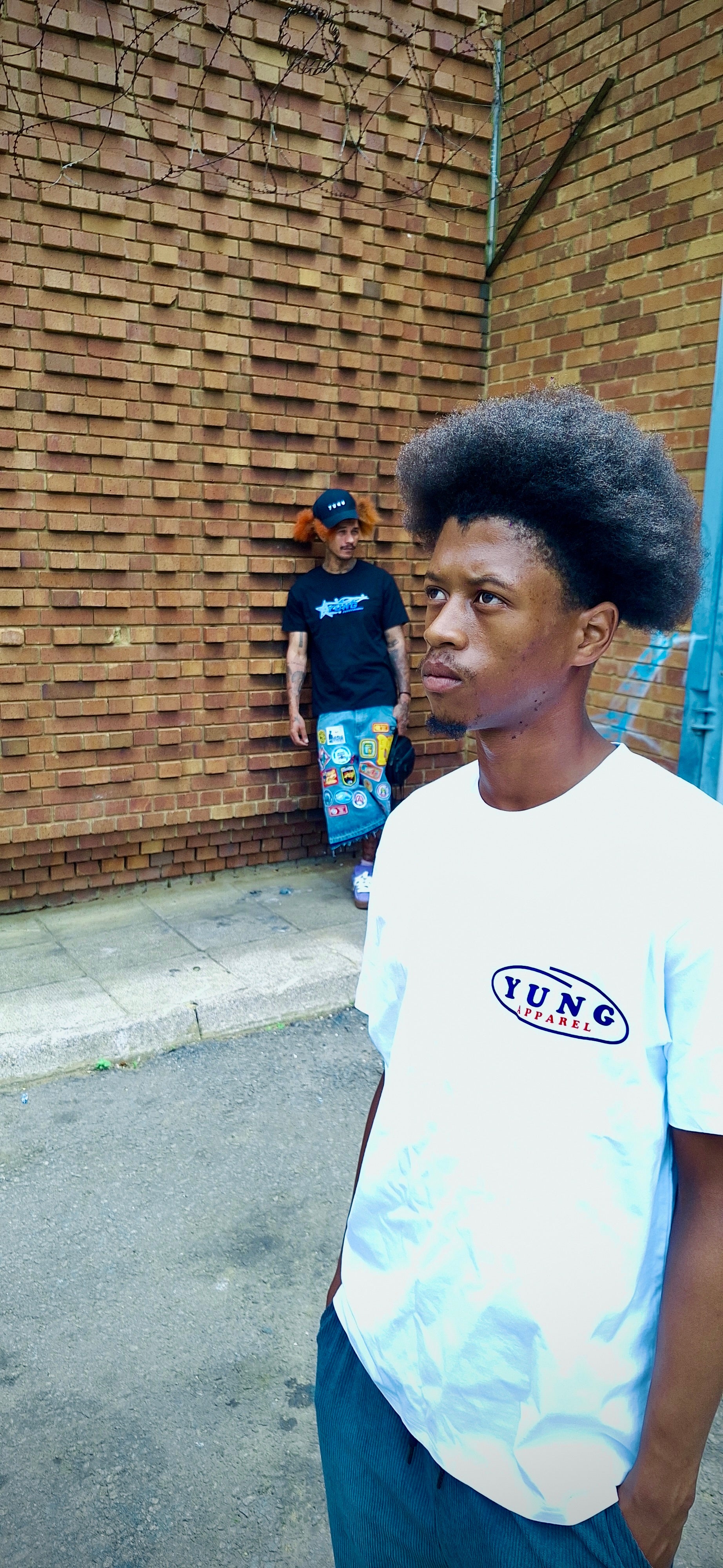 Two people wearing a YUNG black and white t-shirt with a logo, standing in front of a brick wall.
South Africa