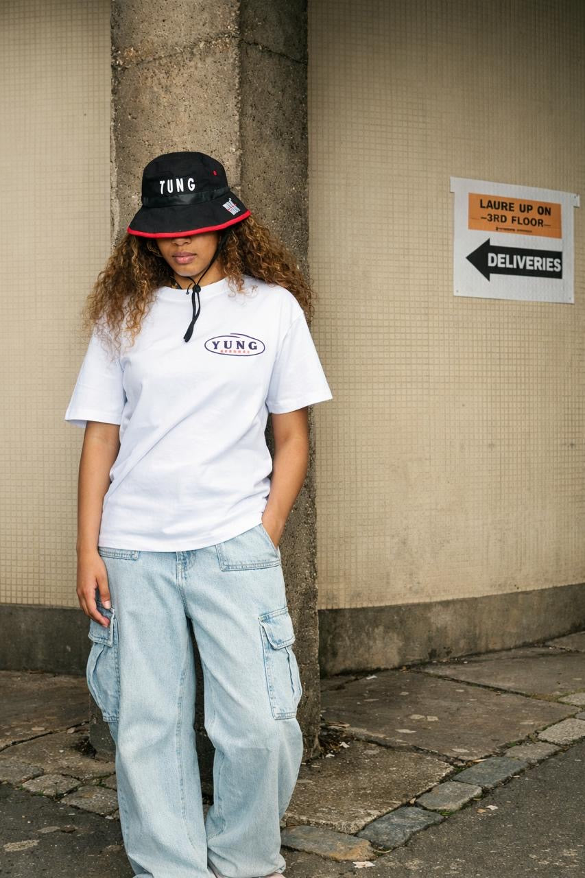 Person wearing a white t-shirt with 'YUNG' logo print, light blue jeans, and a YUNG black bucket hat with red brim, leaning against a wall.
South Africa