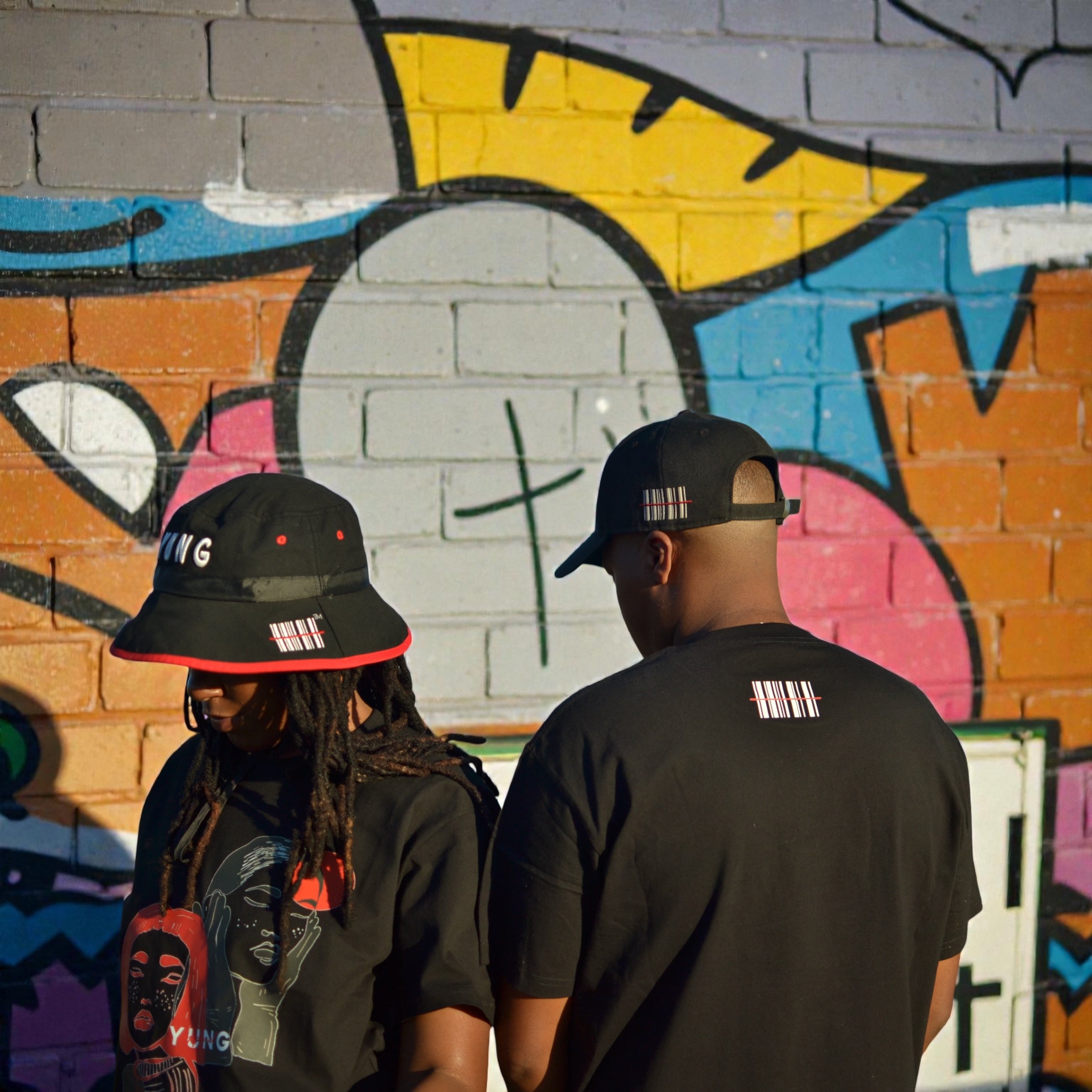 Two people wearing black YUNG bucket hats and baseball cap and YUNG T-shirts in front of a colorful graffiti wall.
South Africa