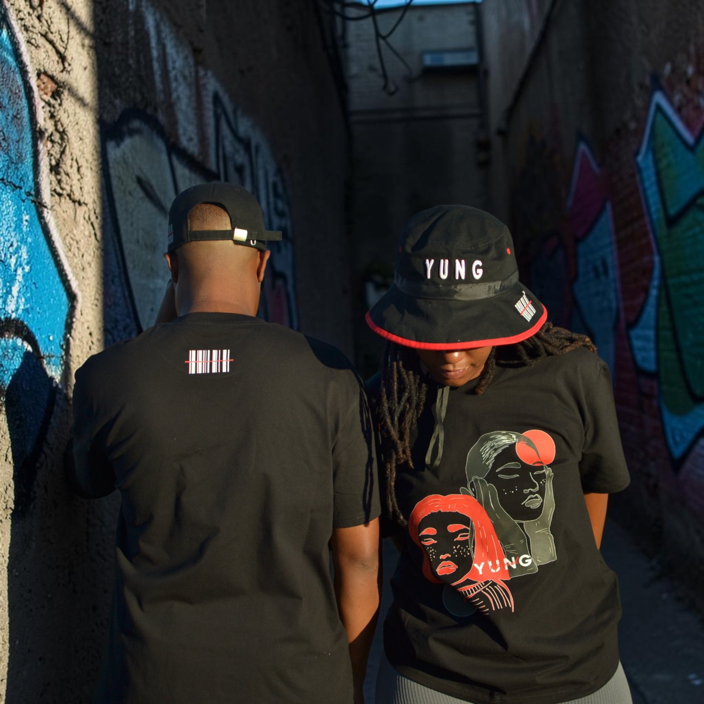 Two people wearing black t-shirts with YUNG graphic designs and YUNG black bucket hat and baseball cap, standing against a graffiti-covered wall.
South Africa