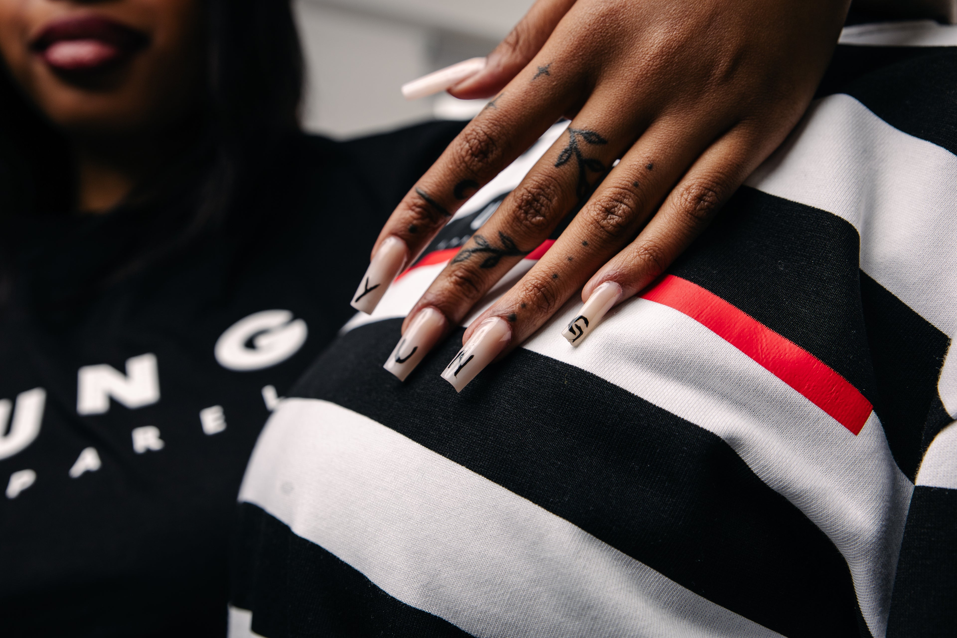 Close-up of a hand with tattoos and  YUNG long nails wearing a black, white, and red striped garment.
Streetwear South Africa